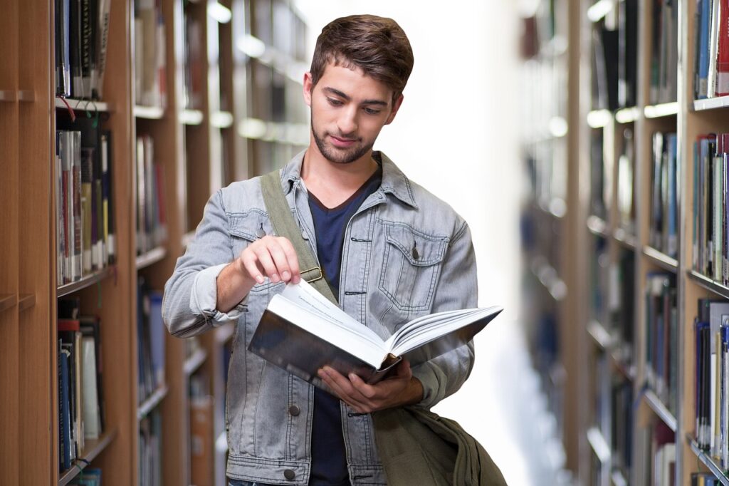 Un joven estudiante con chaqueta de mezclilla y una mochila al hombro está hojeando un libro entre los estantes de una biblioteca universitaria.