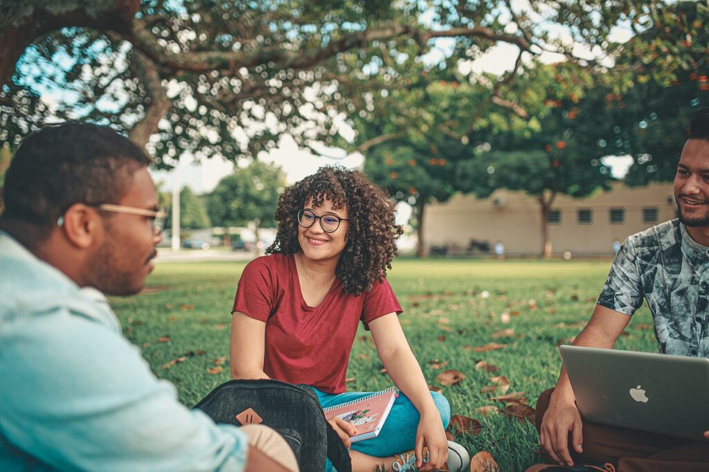 Un grupo de tres estudiantes universitarios sentados en el césped, interactuando alegremente mientras trabajan con un portátil y toman notas, disfrutando de un estudio grupal en un ambiente relajado al aire libre bajo la sombra de un árbol.