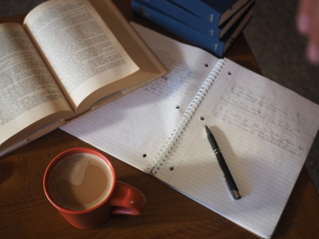 Taza de café con leche al lado de un cuaderno y libros en un ambiente de estudio.